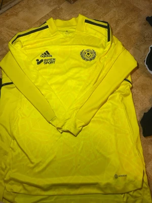 Gul långärmad fotbollströja Adidas - Säljer en gul långärmad fotbollströja från Adidas med svarta detaljer och tryck från Skogås-Trångsunds FF. Tröjan har klubbmärke, Adidas-logga och Intersport-tryck på bröstet. Tillverkad i lätt och ventilerande material, perfekt för träning eller match.