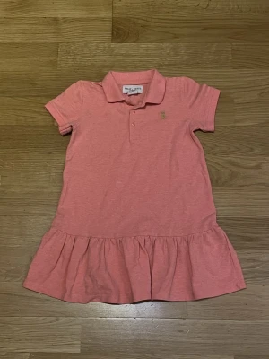 Rosa pikéklänning från Holly & White - Söt rosa pikéklänning med klassisk krage, korta ärmar och volang nedtill. Klänningen har knappar vid halsen och en liten broderad logga på bröstet. .