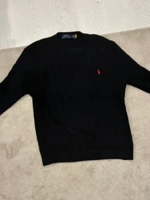 Svart stickad tröja Polo Ralph Lauren - Svart stickad tröja från Polo Ralph Lauren med klassiskt kabelstickat mönster och röd broderad logga på bröstet. Tröjan har rund halsringning och ribbade muddar vid ärmslut och nederkant. Perfekt för en stilren look.