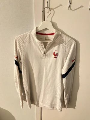 Vit Nike Frankrike träningströja - Snygg vit långärmad träningströja från Nike med Frankrikes landslagsmärke på bröstet. Tröjan har halv dragkedja, blå och röda detaljer på ärmarna samt Nike-logga. Perfekt för fotbollsträning eller sportiga dagar.