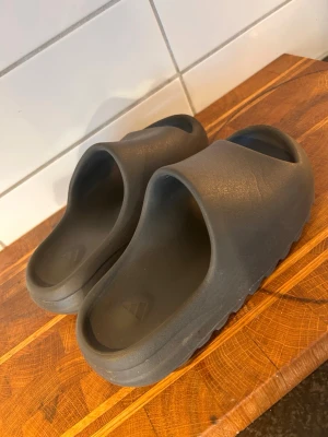 Svarta Yeezy slides - Säljer ett par svarta Adidas Yeezy slides med chunky sula och öppen tå. Tillverkade i ett mjukt och flexibelt syntetmaterial som ger en clean och modern look. Perfekta för chill sommardagar!