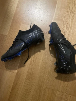 Nike svart/blå fotbollsskor Zoom - Säljer ett par svarta Nike fotbollsskor med blå och vita detaljer samt Zoom-teknologi. Skorna har snörning, låg profil och dobbar för bra grepp på planen. Snygg design med vita linjer och Nike-logga på sidan. Perfekta för dig som vill ha stil och funktion på fotbollsplanen.