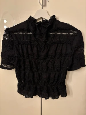 Svart spetsblus - Trendig svart blus i transparent spets med volangdetaljer och hög krage. Kortärmad modell med resår i midjan för en snygg siluett. Stretchig och i strl xs så skulle säga passar xs-m