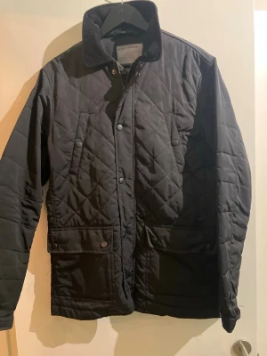 Svart quiltad vindjacka - Snygg svart quiltad vindjacka med krage och tryckknappar och dragtjedja framtill. Jackan har två stora fickor med lock och en stilren, tidlös design. Materialet är lätt och vindtätt, perfekt för blåsiga dagar. Passar dig som gillar klassisk men modern stil.