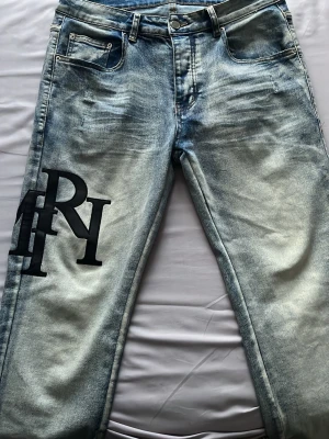 Ljusblåa amiri jeans  - Säljer ett par ljusblå tvättade jeans med stor svart logotyp på vänster lår. Jeansen har klassisk femficksmodell, raka ben och knappgylf. Materialet är denim i bomull och de har en trendig, sliten look. Perfekta för dig som gillar streetwear och vill sticka ut. Säljer den på grund av att jag inte längre använder de. Fick som present.