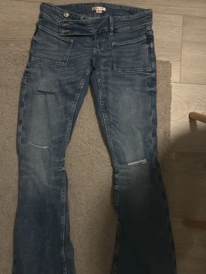 Blå slitna bootcut jeans - Säljer ett par blå bootcut jeans med slitningar på benen och dubbla knappar i midjan. Jeansen har stora fickor fram och en cool tvättad look. Perfekta för dig som gillar en avslappnad och trendig stil.