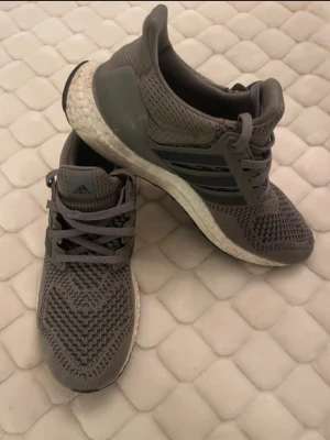 Grå Adidas Ultraboost sneakers - Snygga grå Adidas Ultraboost sneakers med stickad ovandel i mesh och klassiska tre ränder på sidan. Skorna har en vit Boost-sula för extra komfort och gråa snören. Perfekta för dig som gillar sportig och modern stil. Kan gå ner i pris vid snabb affär
