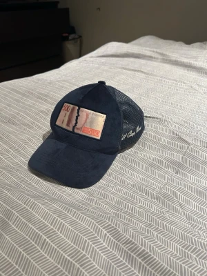 Blå Bruce Lee keps med mesh - Snygg mörkblå keps från Bruce Lee med mesh på sidorna och bak, samt ett patch framtill med motiv av en 500-kronorssedel. Justerbar baktill för perfekt passform. Perfekt för dig som vill sticka ut med en unik accessoar.