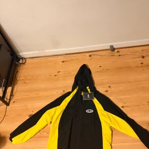 Svart och gul vindjacka från Corteiz - Svart och gul vindjacka från Corteiz med huva och coola kontrastpaneler. Jackan har dragkedja framtill, snörning vid huvan och ett broderat märke på bröstet. Materialet är lätt och syntetiskt, perfekt för att sticka ut med streetstyle-vibe.