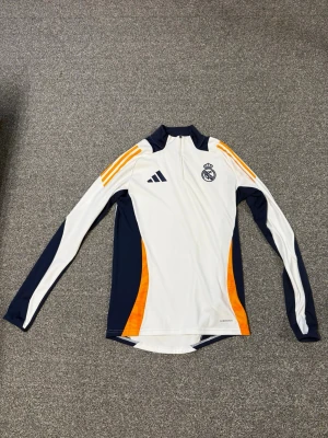 Real Madrid träningströja Adidas - Snygg långärmad träningströja från Adidas med Real Madrid-logga. Tröjan är vit med marinblå och orange detaljer, halv dragkedja framtill och klassiska Adidas-ränder på axlarna. Tillverkad i funktionsmaterial som andas, perfekt för fotbollsträning.