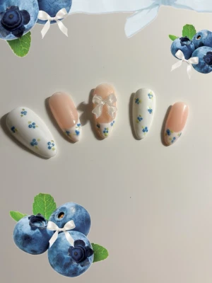 Press-on naglar med blå blommor och rosett - Söta press-on naglar i mandelform med vit och ljusrosa bas, dekorerade med små blå blommor och en 3D-rosett på en nagel. Perfekt för dig som vill ha en gullig och unik nageldesign. Materialet är plast.