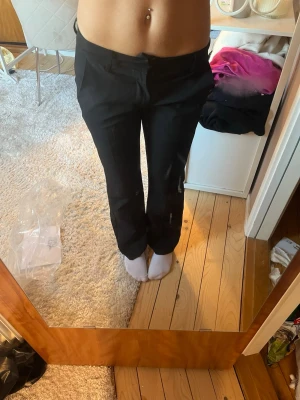 Svarta raka kostymbyxor i petite - Snygga svarta kostymbyxor med rak passform och klassiska fickor bak. De är storlek petite i längden. Byxorna har hällor för skärp och stängs med knapp och dragkedja. Perfekta för en stilren och clean look. Materialet ser ut att vara en mjuk och följsam blandning, troligen polyester eller bomull.