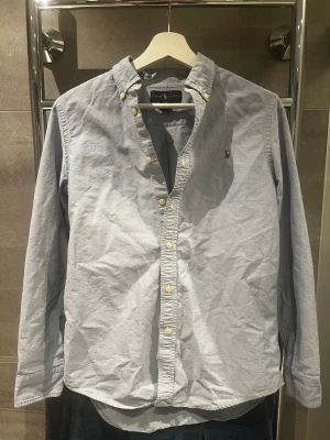 Ljusblå henley från Ralph Lauren - Snygg ljusblå henley-skjorta från Ralph Lauren med klassisk button-down krage och guldfärgade knappar. Skjortan har lång ärm och den ikoniska broderade loggan på bröstet. Tillverkad i mjuk bomull för en bekväm känsla. Nypris: runt 1200kr. Storlek S. Skriv vid frågor!