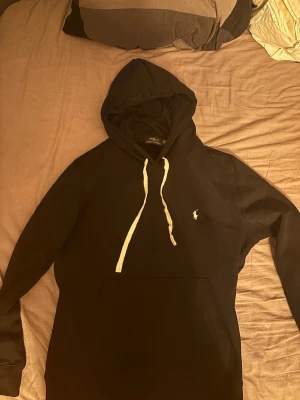 Svart hoodie från Polo Ralph Lauren - Svart hoodie från Polo Ralph Lauren med vit dragsko och klassisk logga broderad på bröstet. Hoodien har en stor ficka framtill och en skön huva. Perfekt för en chill och stilren look.