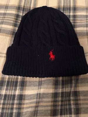 Mörkblå kabelstickad mössa Polo Ralph Lauren - Snygg mörkblå kabelstickad mössa från Polo Ralph Lauren med uppvikt kant och klassisk röd logobrodyr framtill. Tillverkad i mjukt garn som håller dig varm och ger en clean look. Perfekt accessoar för kalla dagar.