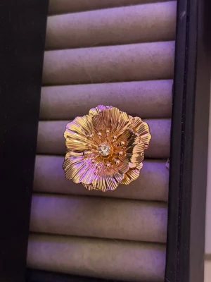 Guldfärgad blomring med kristall - Unik ring i guldfärgad metall formad som en stor blomma med detaljerade kronblad och en gnistrande klar sten i mitten. Perfekt för dig som vill sticka ut med ett statement-smycke. Ringen har en elegant och konstnärlig design som drar blickarna till sig.