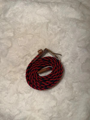 Randigt rött och svart bälte - Snyggt bälte med randigt mönster i rött och svart. Spännet är i metall och detaljerna i brunt skinn. Perfekt accessoar för att lyfta din outfit med lite färg och stil.