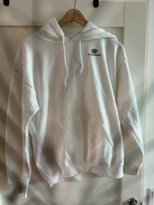 Vit hoodie från Flowwear med tryck - Säljer en vit hoodie från Flowwear med liten logga på bröstet och stort rött joker-tryck på ryggen. Hoodien har huva med dragsko och en stor magficka. Tillverkad i mjukt material, perfekt för en chill och avslappnad stil.