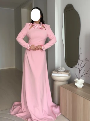 Ljusrosa långklänning med blommiga detaljer - Elegant ljusrosa långklänning med långa ärmar och blommiga detaljer vid krage och ärmslut. Klänningen har hög hals och en smal passform upptill, samt en lätt utsvängd kjol. Perfekt för speciella tillfällen.