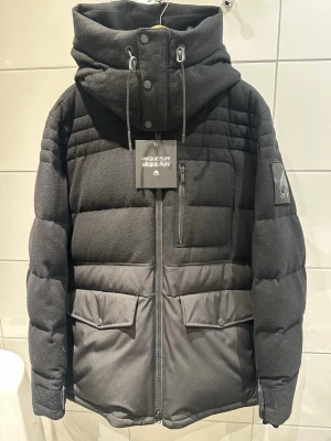 Svart pufferjacka från Moose Knuckles - Säljer en svart pufferjacka från Moose Knuckles med stor huva, dragkedja framtill och flera fickor. Jackan har en cool patch på ärmen och detaljer i metall. Materialet är tjockt och quiltat, perfekt för kalla vinterdagar. Snygg och modern design med hög krage och justerbar huva.