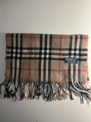 Beige rutig halsduk från Burberrys - Klassisk beige halsduk från Burberrys i 100% kashmir. Den har det ikoniska rutmönstret i svart, vitt och rött samt fransar i båda ändar. Perfekt accessoar för att lyfta din outfit med en tidlös brittisk stil.