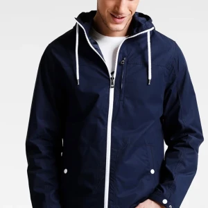 Mörkblå vindjacka med huva - Snygg mörkblå vindjacka med vit dragkedja och vita snören i huvan. Jackan har två fickor med knappar och en sportig look. Perfekt för blåsiga dagar och enkel att matcha med olika outfits.