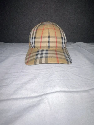 Burberry keps med klassiskt rutmönster - Snygg keps från Burberry i deras ikoniska beigea rutmönster med svarta, vita och röda ränder. Kepsen har böjd skärm och justerbar passform. Perfekt accessoar för en trendig och stilren look.