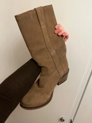 Beige mocka cowboy boots - Snygga cowboy boots i beige mocka med klassisk hög skaft och markerad klack. Bootsens stilrena design och mjuka material gör dem till ett statement-piece för dig som gillar western-inspirerat mode.