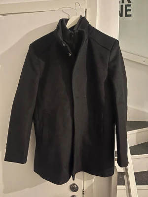 Svart klassisk rock med ståkrage - Snygg svart rock med ståkrage och dold knäppning. Rocken har raka linjer, två sidofickor och dekorativa knappar vid ärmslutet. Perfekt för dig som vill vara stilren. Original pris 1200kr