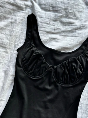 Svart tight kort klänning - Säljer en svart tight kort klänning med breda axelband och rynkade detaljer vid bysten. Klänningen har en enkel, stilren design och är perfekt för dig som gillar minimalistisk stil. Materialet känns mjukt och stretchigt.