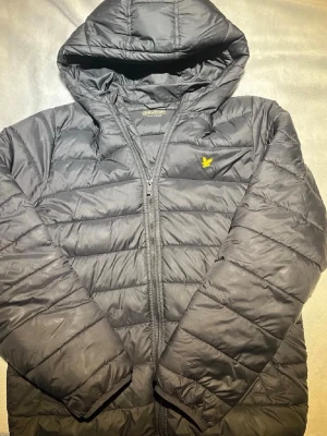 Svart dunjacka från Lyle & Scott - Snygg svart dunjacka från Lyle & Scott med gul logga på bröstet. Jackan har huva, dragkedja framtill och två snedställda fickor. Quiltad design med horisontella sömmar och lätt glansigt material. Perfekt för kalla vinterdagar.