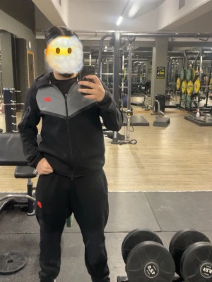Svart träningsset från Li-Ning - Säljer ett svart träningsset från Li-Ning med matchande zip-hoodie och byxor. Hoodien har grå panel över bröstet och röd logga på vänster sida. Byxorna är svarta med röd logga på benet. Perfekt för gymmet eller sportaktiviteter.