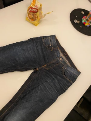 Mörkblå jeans från Nudie Jeans - Säljer ett par mörkblå jeans från Nudie Jeans med klassiska kontrastsömmar och snygga detaljer på bakfickorna. Jeansen har rak passform och är tillverkade i slitstarkt bomullsjeansmaterial. Perfekta för dig som gillar stilrena och tidlösa jeans.