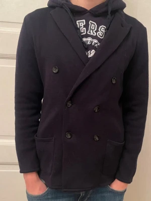 Mörkblå  kavaj från John Henric  - Stilren mörkblå kavaj med dubbelknäppning och breda slag. Merino ull Två framfickor och mjukt, stickat material som ger en avslappnad men ändå dressad look. Passar perfekt till jeans eller chinos för en modern stil.