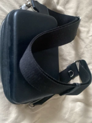 Svart boxig handväska med bred rem - Cool svart handväska i boxig form med bred, justerbar axelrem. Väskan har ett hårt yttre skal i läderimitation och en silverfärgad dragkedja. Perfekt för dig som gillar minimalistisk och stilren design.