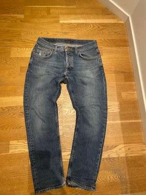 Blå jeans från Nudie Jeans - Säljer ett par klassiska blå jeans från Nudie Jeans med fem fickor och orangea sömmar. Jeansen har en rak passform och snygga slitningar. Tillverkade i denim med en cool tvättad look.