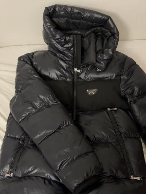 Svart pufferjacka från Alessandro - Snygg svart pufferjacka från Alessandro med glansig finish och stora vadderade partier. Jackan har huva, två snedställda dragkedjefickor och en cool logga på bröstet. Perfekt för kalla vinterdagar och riktigt trendig look.