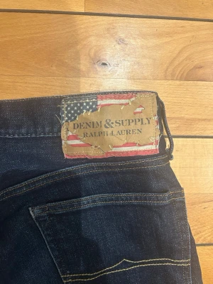Denim & Supply Ralph Lauren jeans - Snygga mörkblå jeans från Ralph Lauren, Denim & Supply-kollektionen. Klassisk femficksmodell med smal passform och cool detalj med amerikansk flagga på läderpatchen bak. Perfekta till en avslappnad streetstil. Ny pris 1999kr. Priset är inte fast