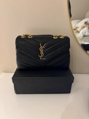 Svart YSL axelväska i läder - Snygg svart axelväska från YSL i quiltat läder med guldiga kedjor och ikonisk YSL-logga framtill. Väskan har en stilren, rektangulär form och detaljer i guldmetall. Perfekt för dig som vill ha en lyxig och trendig accessoar.