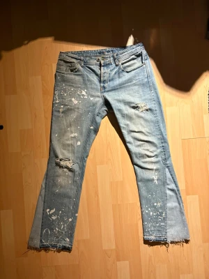Ljusblå slitna jeans med färgstänk - Ett par ljusblå vintage NN07 jeans som är reworked med rå och bredare benöppning samt en paint-splatter design. Flaws vid grenen samt en reva vid benöppningen. Överlag jävligt feta jeans.