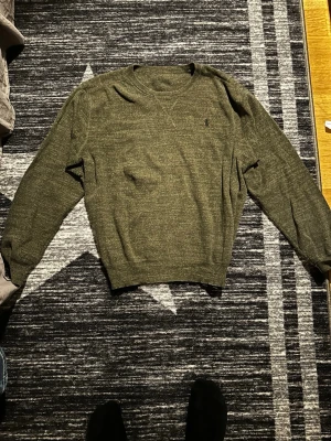 Olivgrön stickad tröja från Polo Ralph Lauren - Snygg olivgrön stickad tröja från Polo Ralph Lauren med klassisk broderad logga på bröstet. Tröjan har rund halsringning, långa ärmar och ribbade muddar vid ärmslut och nederkant. Perfekt för lager på lager och enkel att matcha. Tyvärr så e lappen av men äkta såklart om man kollar på hästen