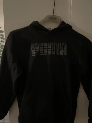 Svart hoodie från Puma med tryck - Svart hoodie från Puma med stort grafiskt PUMA-tryck på bröstet. Klassisk modell med huva och känguruficka framtill. Perfekt för en sportig och avslappnad stil. Tillverkad i mjukt material för extra komfort.