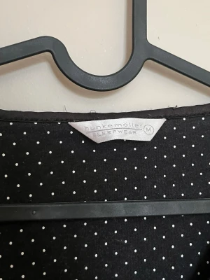Svart nattlinne från Hunkemöller - Mysigt svart nattlinne från Hunkemöller med vita små prickar på insidan av halsringningen. Långärmad modell med rund hals och små rosetter vid nederkanten. Perfekt för chillkvällar hemma.
