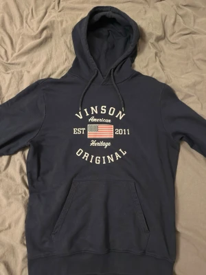 Mörkblå hoodie Vinson Original - Mörkblå hoodie från Vinson Original med tryck framtill som inkluderar amerikansk flagga och texten 'American Heritage EST 2011'. Klassisk känguruficka och justerbar huva med dragsko. Perfekt för en avslappnad stil.