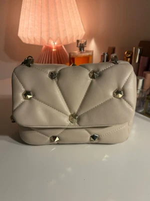Väska med nitar  - Snygg väska med nitar, liknar valentino väskan. Krämvit/beige med guld detaljer. Perfekt skick bort sätt från den pytte lilla repan på insidan (syns på bild 6) ❣️