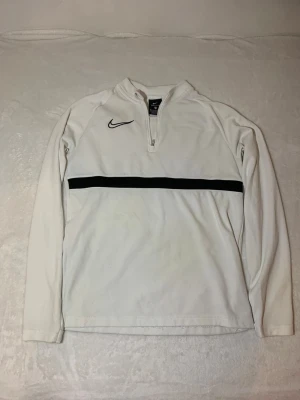 Vit Nike Dri-FIT träningströja - Vit långärmad träningströja från Nike med svart logga på bröstet och en svart horisontell rand över bröstet. Tröjan har halv dragkedja och är tillverkad i Dri-FIT-material som andas. Perfekt för sport och träning.