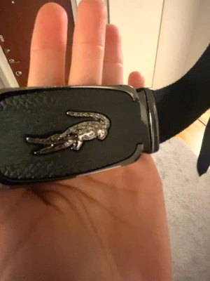 Svart bälte från Lacoste med metallspänne - Snyggt svart bälte från Lacoste med ett metallspänne som har den klassiska krokodilloggan. Bältet har en stilren design och passar perfekt till jeans eller byxor. Materialet känns slitstarkt och spännet ger en exklusiv touch.