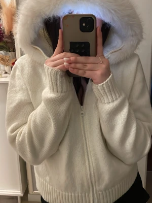Vit stickad hoodie med fuskpäls - Mysig vit stickad hoodie med dragkedja och stor huva kantad med fluffig fuskpäls. Tröjan har ribbstickade muddar vid ärmslut och nederkant. Perfekt för kalla dagar när du vill vara både varm och snygg.pris kan diskuteras