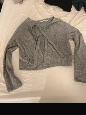 Grå croppad tröja med knytband - Säljer en grå croppad tröja med långa ärmar och snygga knytband vid halsen. Tröjan är mjuk och har en enkel, stilren design som passar perfekt till jeans eller kjol. Materialet känns stretchigt och bekvämt.