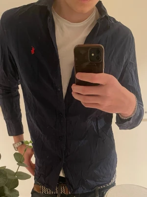 Mörkblå skjorta från Ralph Lauren - Säljer en mörkblå slim fit stretch oxford-skjorta från Ralph Lauren med klassisk krage och röd broderad logga på bröstet. Skjortan har långa ärmar och knäppning framtill. Perfekt för en stilren och avslappnad look.